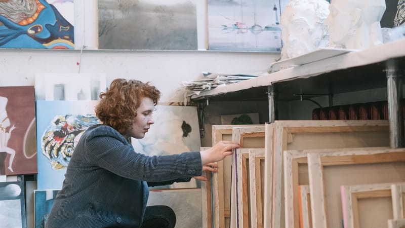 Une artiste concentrée organise des toiles dans un studio d'art en désordre rempli d'œuvres d'art diversifiées.