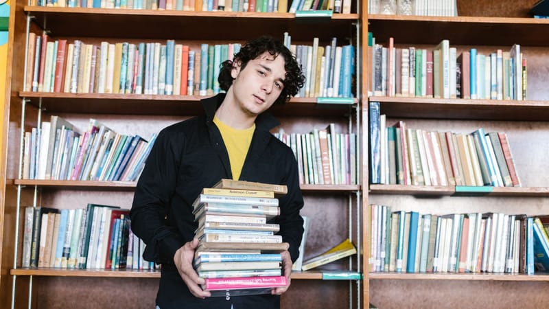 Un jeune homme tenant une pile de livres dans une bibliothèque, symbolisant l'éducation et l'apprentissage.