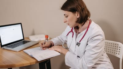 Une médecin portant une blouse d'hôpital écrivant une prescription dans un bureau clinique avec un ordinateur portable sur la table.