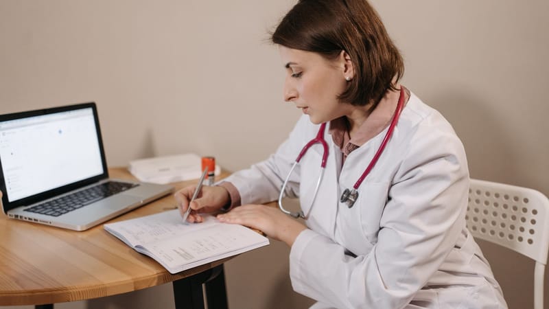 Une médecin portant une blouse d'hôpital écrivant une prescription dans un bureau clinique avec un ordinateur portable sur la table.