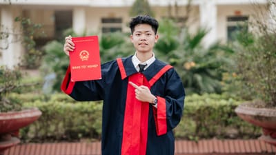 Jeune diplômé en robe à bord rouge tenant fièrement son diplôme à l'extérieur.