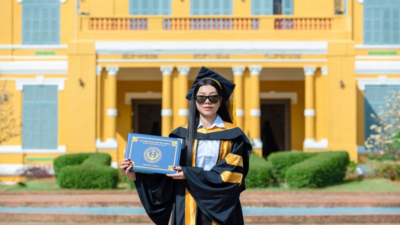 Un diplômé, coiffé d'une toque et vêtu d'une robe de cérémonie, tient son diplôme devant un bâtiment jaune historique.