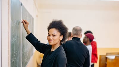 Une étudiante écrivant avec confiance sur un tableau noir dans une salle de classe animée avec ses camarades.