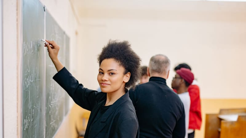 Une étudiante écrivant avec confiance sur un tableau noir dans une salle de classe animée avec ses camarades.