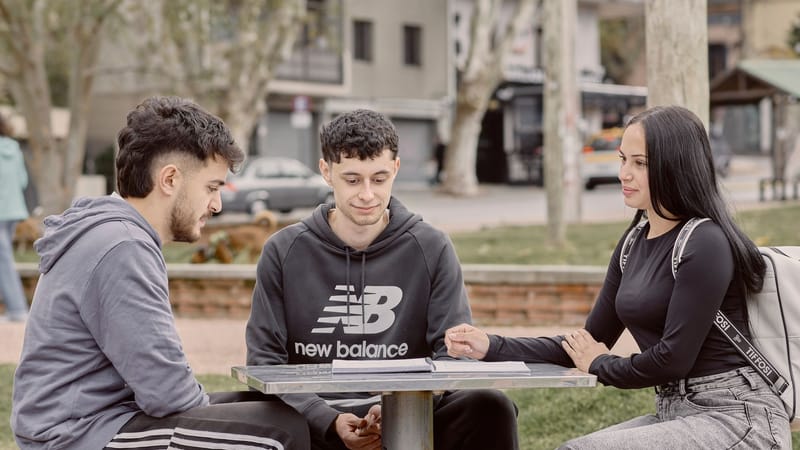 Trois jeunes adultes étudient ensemble en plein air, attablés à une table de pique-nique dans un parc uruguayen, illustrant ainsi leurs interactions sociales et leur collaboration.
