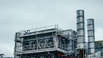 Vue extérieure d'une usine industrielle avec des cheminées métalliques et un réseau de tuyauterie complexe sous un ciel nuageux.
