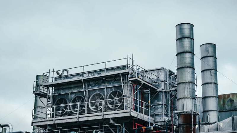 Vue extérieure d'une usine industrielle avec des cheminées métalliques et un réseau de tuyauterie complexe sous un ciel nuageux.