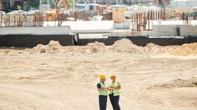 Deux ouvriers discutent des plans sur un chantier de construction sablonneux, portant des équipements de sécurité.