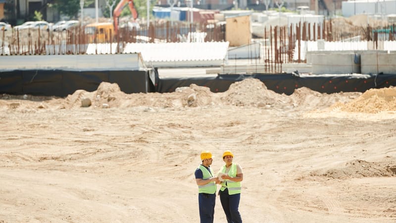 Deux ouvriers discutent des plans sur un chantier de construction sablonneux, portant des équipements de sécurité.