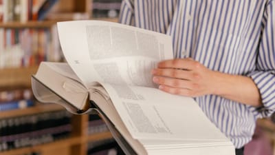 Gros plan sur une personne lisant un grand livre ouvert dans un cadre de bibliothèque avec des étagères à livres en arrière-plan.