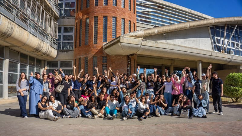 Grand groupe de jeunes adultes divers célébrant devant un bâtiment de campus moderne par une journée ensoleillée.