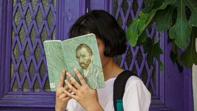 Une jeune femme lit un livre d'art de Van Gogh devant une porte violette, partiellement cachée par des feuilles de figuier.