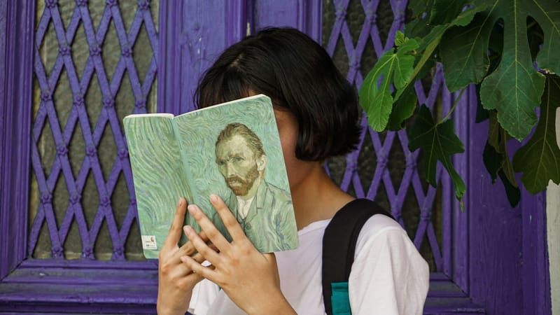 Une jeune femme lit un livre d'art de Van Gogh devant une porte violette, partiellement cachée par des feuilles de figuier.