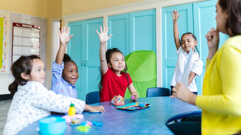 Enfants levant les mains avec enthousiasme dans une salle de classe colorée, engagés dans l'apprentissage.