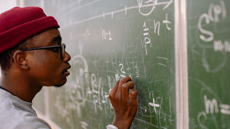 Un jeune homme portant des lunettes et un bonnet rouge résolvant des problèmes de mathématiques sur un tableau noir.