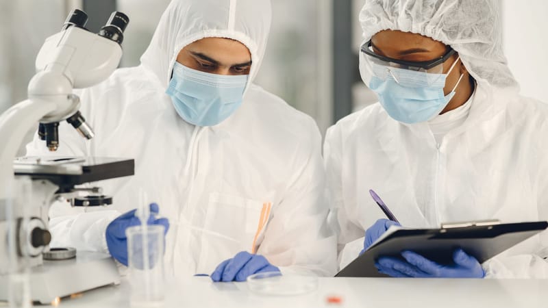 Deux scientifiques en combinaisons de protection travaillant avec un microscope et des notes dans un environnement de laboratoire.