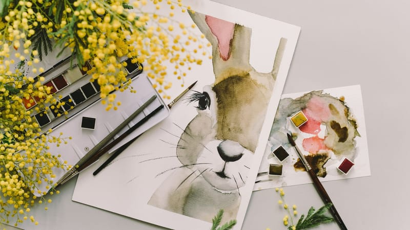 Une peinture à l'aquarelle d'un lapin entouré de fleurs de mimosa et de fournitures artistiques.