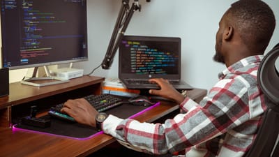 Un homme africain codant sur un ordinateur de bureau et un ordinateur portable dans un bureau à Nairobi, mettant en valeur la technologie moderne.