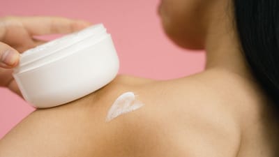 Femme appliquant de la crème de soin sur l'épaule sur un fond rose.