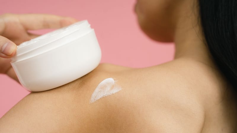 Femme appliquant de la crème de soin sur l'épaule sur un fond rose.