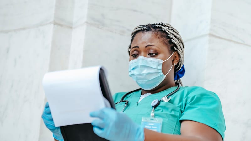 Infirmière afro-américaine portant un masque, révisant des documents dans un cadre hospitalier.