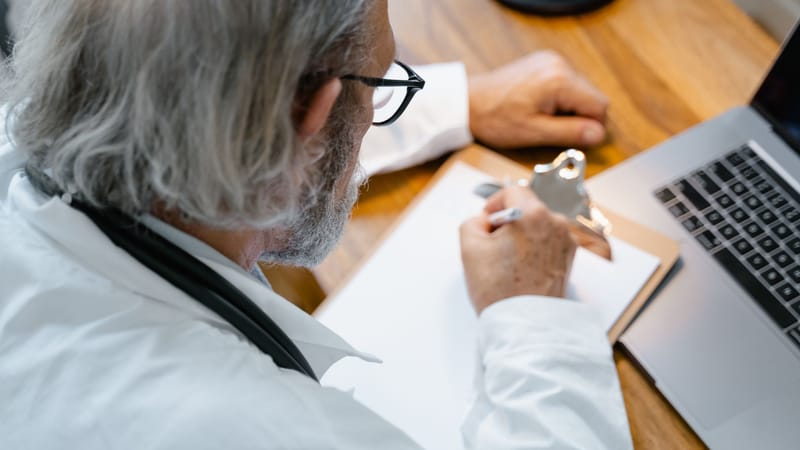 Un médecin âgé en blouse blanche prenant des notes sur un cahier à un bureau avec un ordinateur portable.