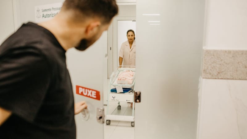 Un homme ouvre une porte pour voir son nouveau-né dans une salle d'hôpital assisté par une infirmière.