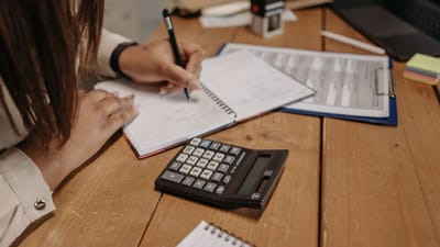 Femme prenant des notes avec une calculatrice sur un bureau en bois, concentrée sur son travail et sa comptabilité.