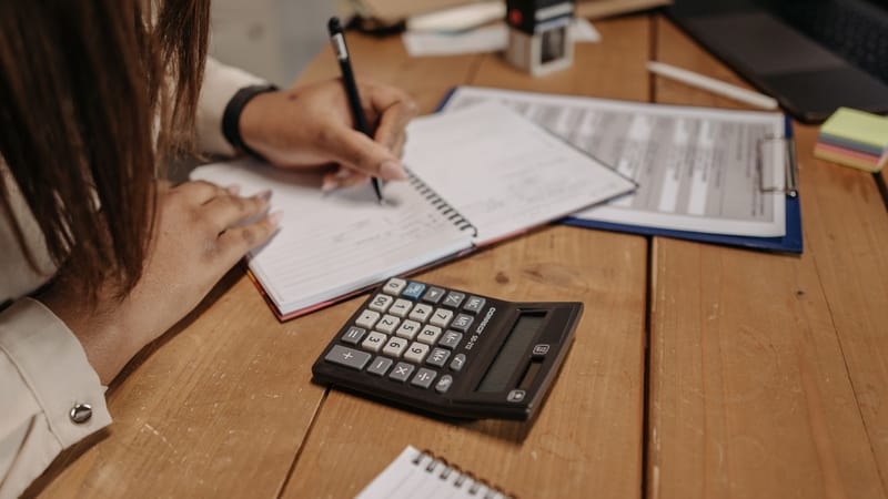 Femme prenant des notes avec une calculatrice sur un bureau en bois, concentrée sur son travail et sa comptabilité.