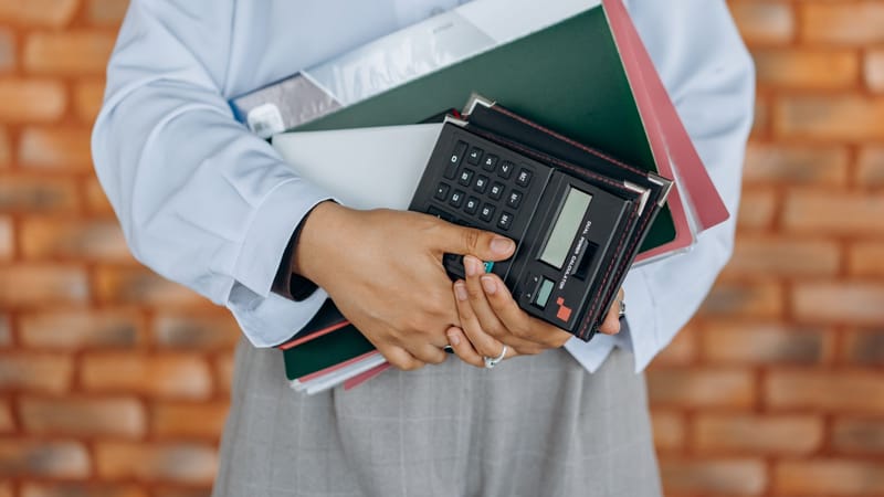 Gros plan sur une personne tenant une calculatrice et des dossiers sur fond de mur en briques.