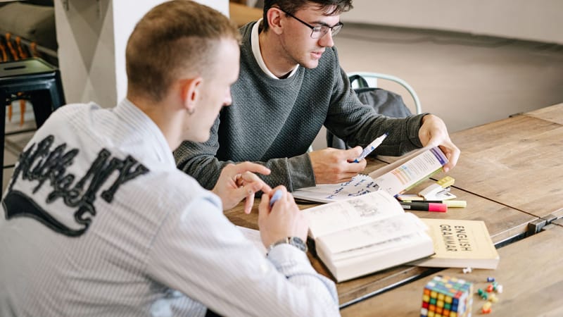 Deux jeunes hommes étudiant ensemble, concentrés sur des livres de grammaire anglaise dans un cadre intérieur moderne.