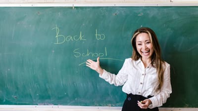 Enseignant souriant dans une salle de classe pointant le message de rentrée scolaire sur un tableau noir.