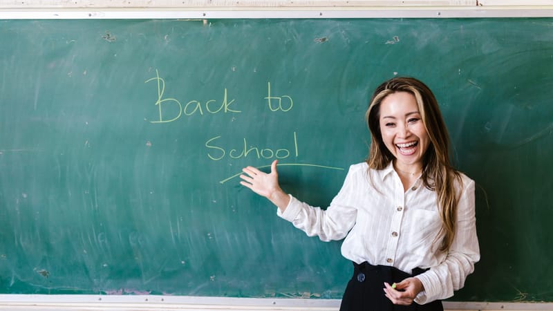 Enseignant souriant dans une salle de classe pointant le message de rentrée scolaire sur un tableau noir.