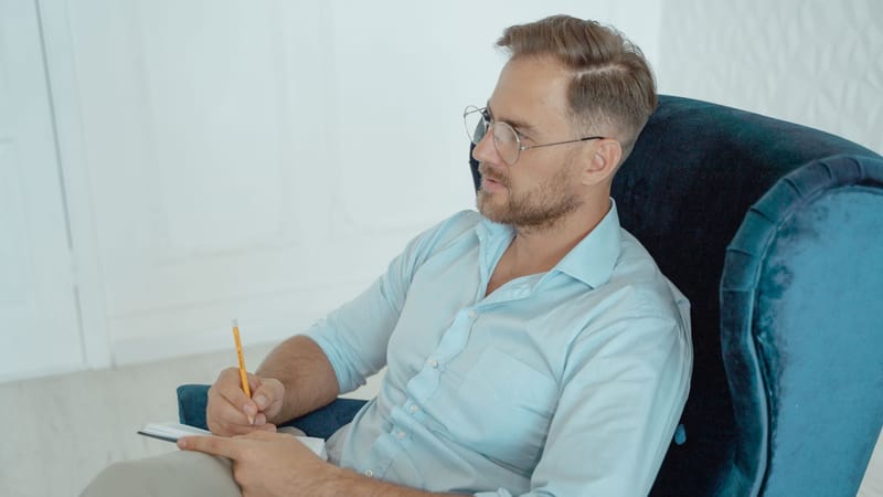 Un thérapeute en séance, portant des lunettes et prenant des notes tout en étant assis sur un fauteuil.