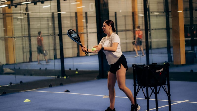 Une jeune femme tenant une raquette et une balle sur un court de paddle-tennis intérieur, prête à servir.