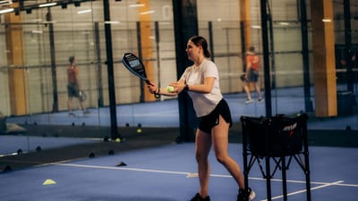 Une jeune femme tenant une raquette et une balle sur un court de paddle-tennis intérieur, prête à servir.
