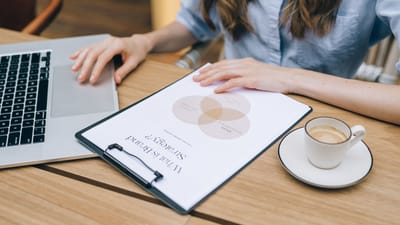 Une personne examine un document de stratégie de marque sur un clipboard tout en utilisant un ordinateur portable dans un environnement de bureau.
