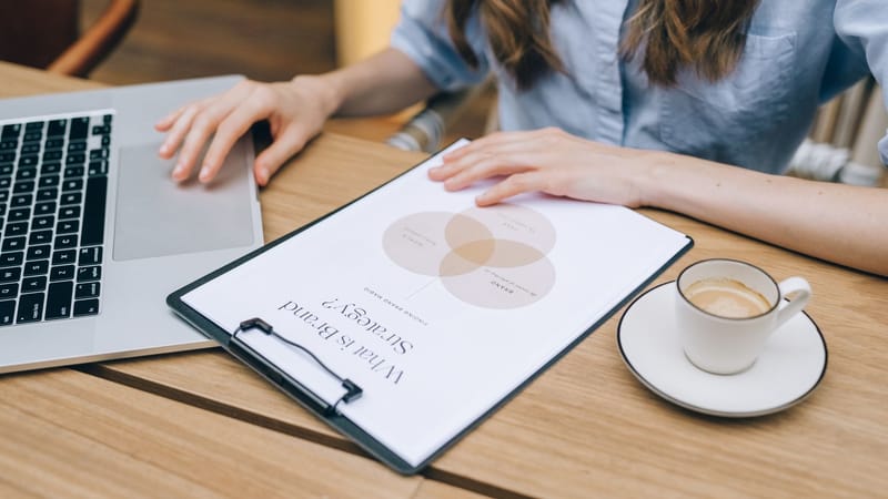 Une personne examine un document de stratégie de marque sur un clipboard tout en utilisant un ordinateur portable dans un environnement de bureau.