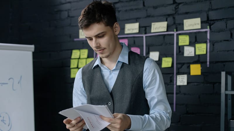 Un jeune homme examinant des documents dans un bureau avec un mur en briques noires et un fond de notes autocollantes.