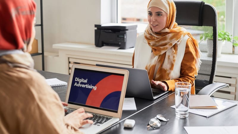 Deux femmes en hijab travaillant sur des ordinateurs portables dans un bureau discutant de la publicité numérique.