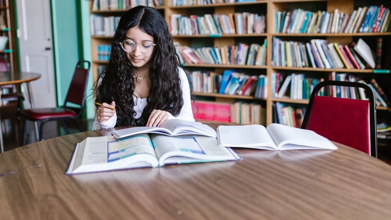 Une jeune femme étudie à une table de bibliothèque entourée de rayonnages, concentrée sur son travail.