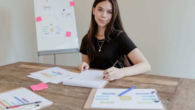 Jeune femme aux cheveux longs étudiant à un bureau avec des documents et un tableau blanc.