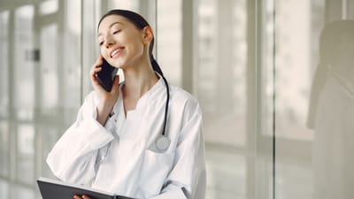 Médecin féminin amical en blouse blanche parlant au téléphone portable, tenant un carnet dans une clinique.