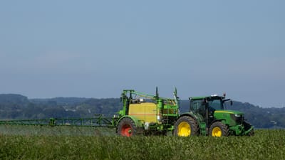 Tracteur vert pulvérisant de l'engrais sur un vaste champ agricole sous un ciel bleu clair.