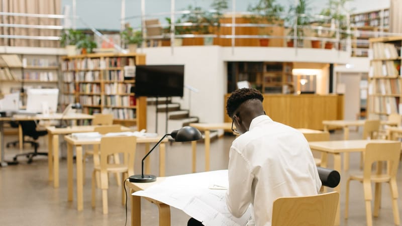 Un étudiant étudie à un bureau dans une bibliothèque moderne avec des étagères et un espace ouvert.