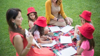 Enfants et enseignant assis sur une couverture dans un parc, engagés dans des activités artistiques.
