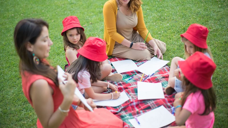 Enfants et enseignant assis sur une couverture dans un parc, engagés dans des activités artistiques.