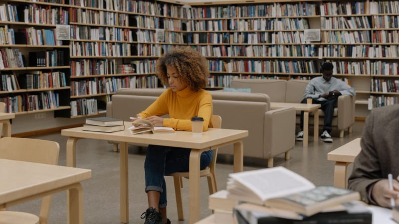 Étudiants étudiant dans une bibliothèque universitaire entourée d'étagères et de tables.