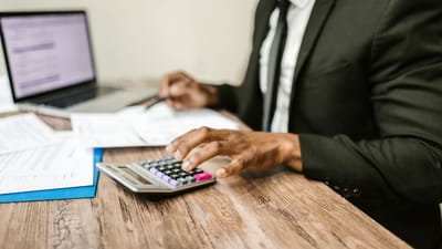 Un homme d'affaires en costume noir utilisant une calculatrice à un bureau avec des documents financiers et un ordinateur portable.