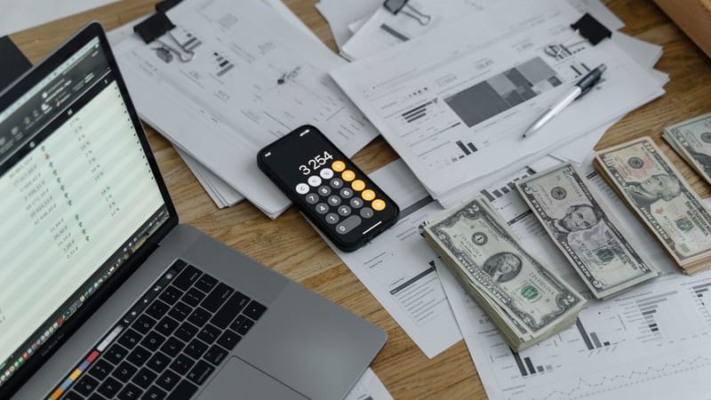 Un bureau avec des documents financiers, de la monnaie, un ordinateur portable et un calculateur de téléphone.